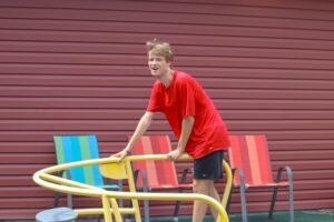 A young person in a red shirt is playfully leaning on a yellow circular structure, with colorful chairs in the background.