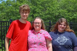 Three young people stand together outdoors, smiling. They are dressed casually, with greenery in the background.