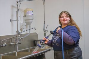 A person stands at a sink, holding a hose with a nozzle, smiling while cleaning. The setting appears to be a kitchen or food preparation area.